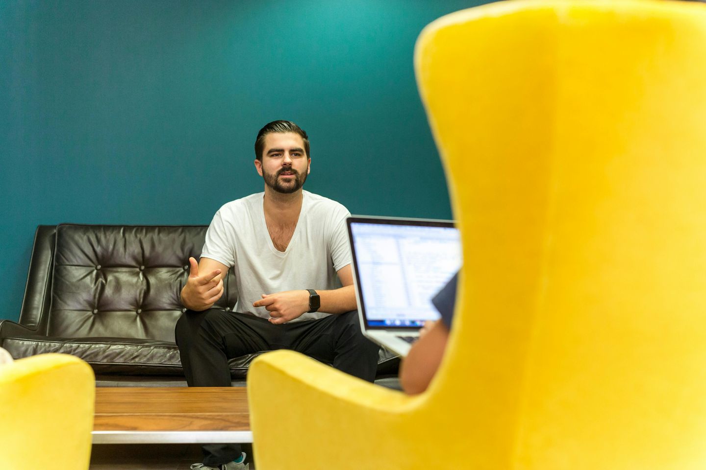 Leadership coach conducting a private coaching session, sitting casually on a leather couch in a modern office with teal walls and yellow chairs, creating an approachable professional environment